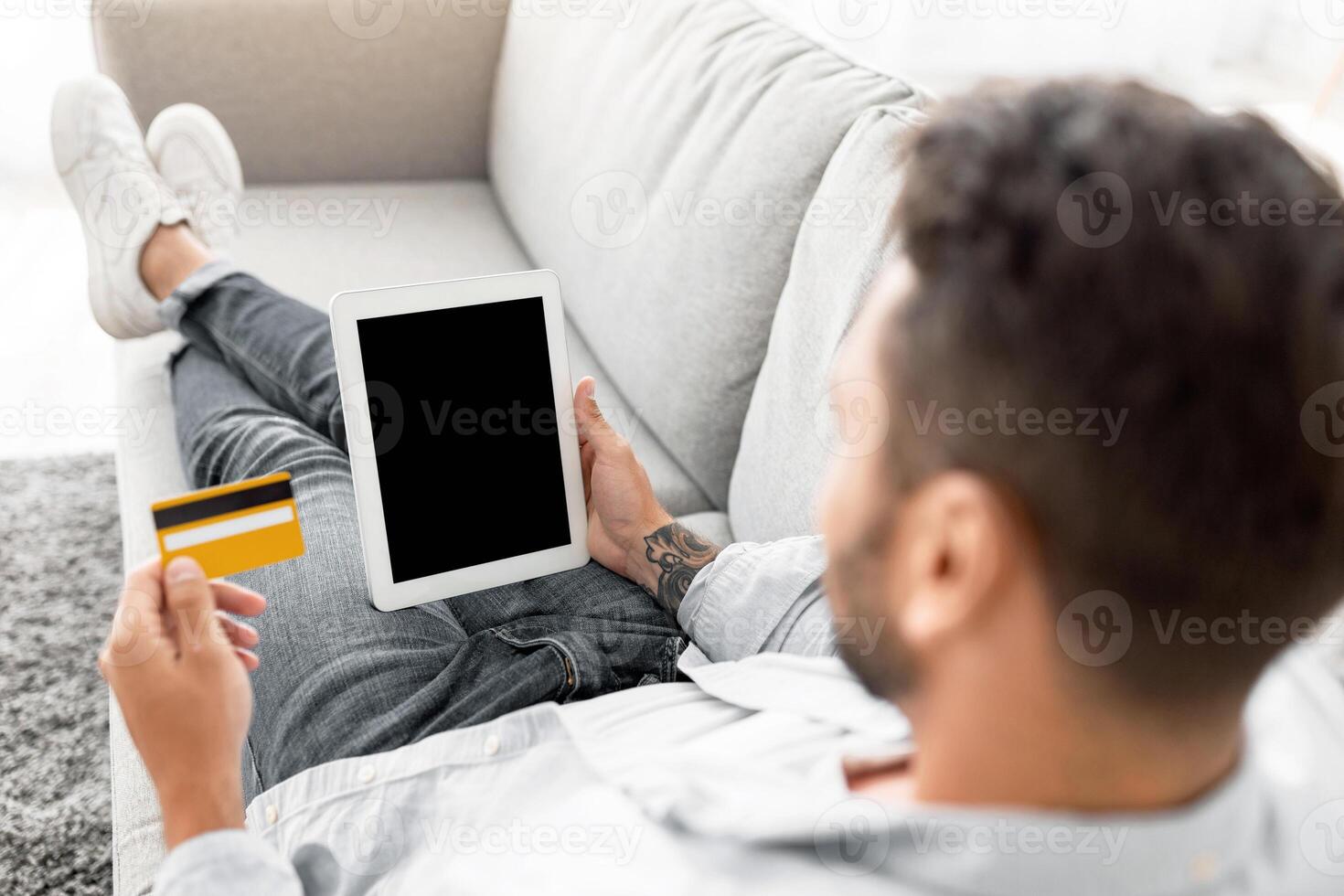 Easy payments, e-commerce, online delivery concept. Young man using tablet pc and credit card, buying goods in internet store, sitting on sofa at home. Mockup for shopping app or website design photo