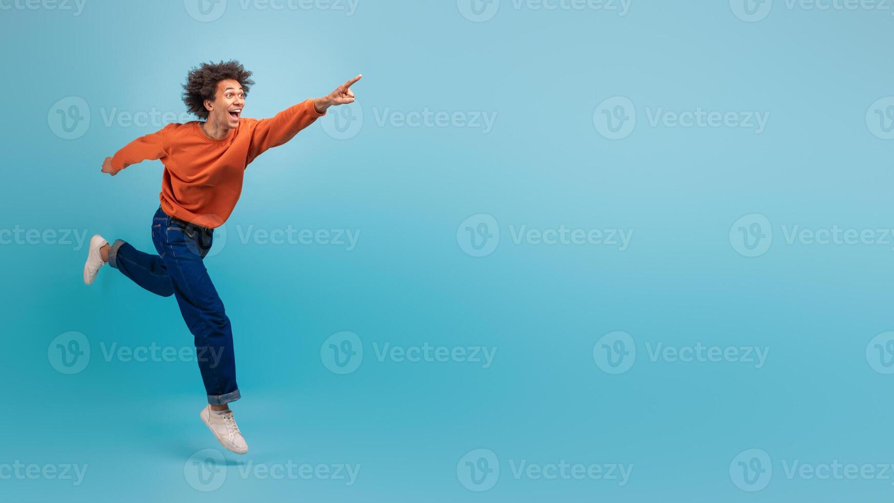 A young African American man with curly hair is energetically running and pointing at copy space, dressed in a casual orange sweatshirt and jeans. photo