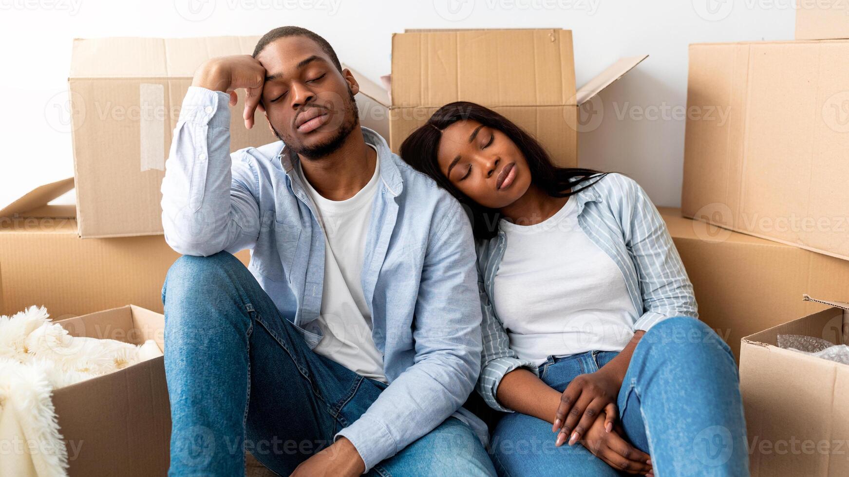 Real estate and renting issues concept. Sleepy african american couple sitting among cardboard boxes, being tired on moving day. Exhausted spouses taking break in unpacking things photo