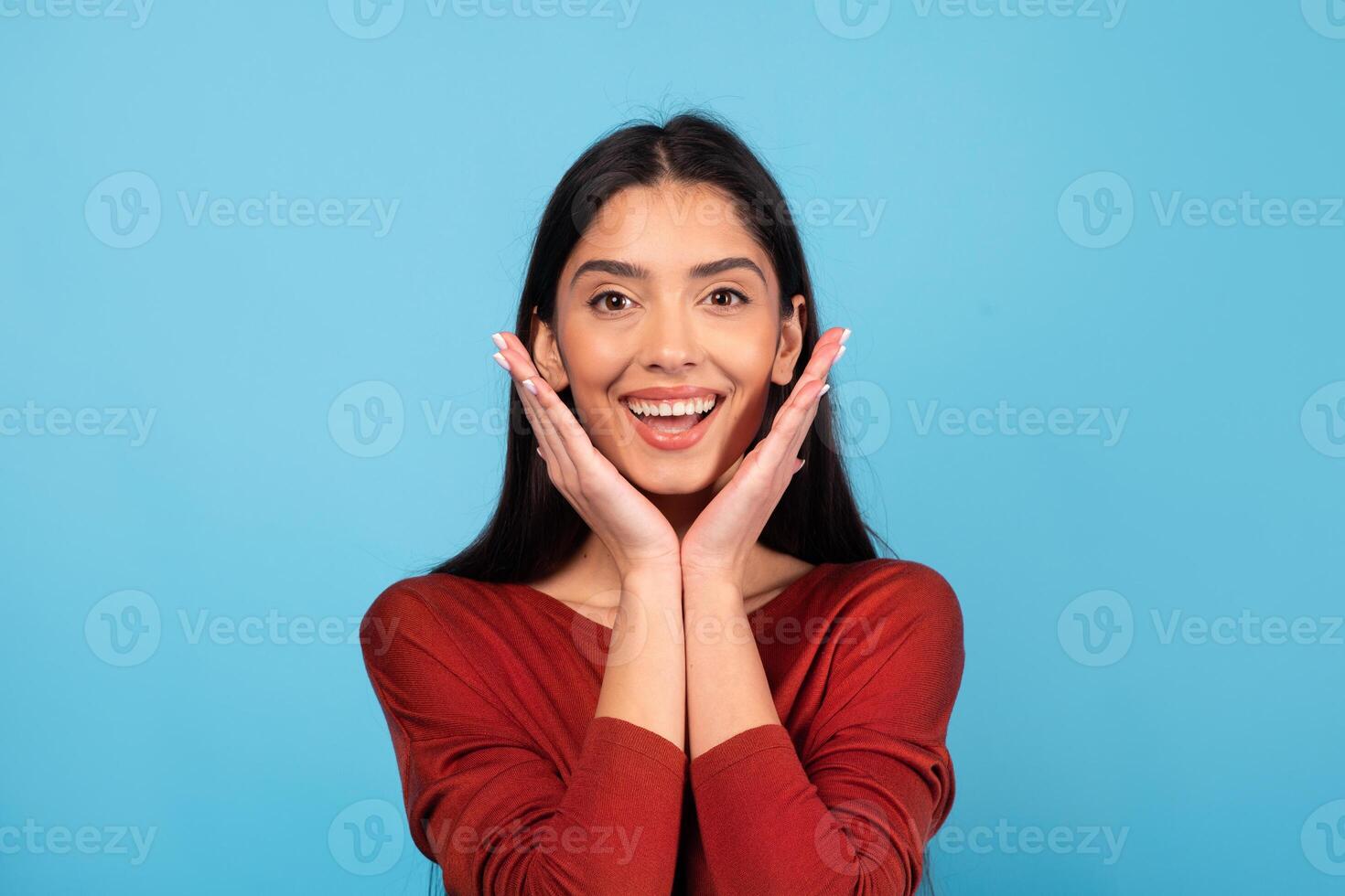 A woman with long brunette hair displays a joyful expression while resting her hands on her cheeks. She is wearing a red sweater and stands in front of a bright blue background, radiating positivity. photo