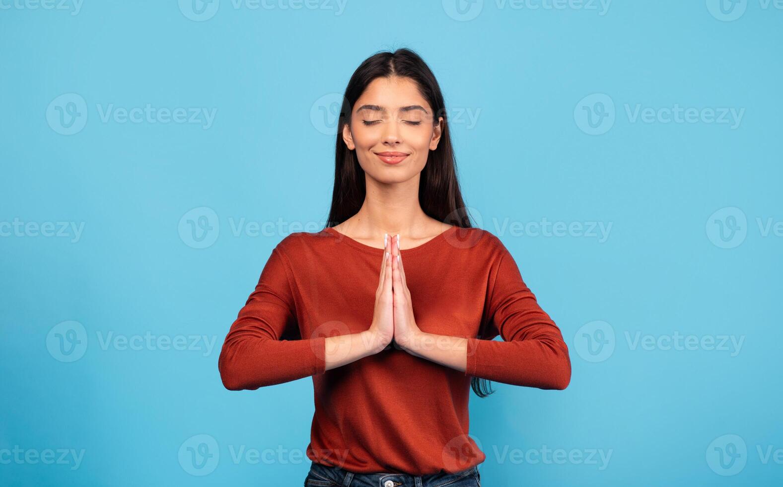 A young Middle Eastern woman displays a peaceful expression as she practices mindfulness. She stands against a vibrant blue background, hands pressed together in prayer position. photo