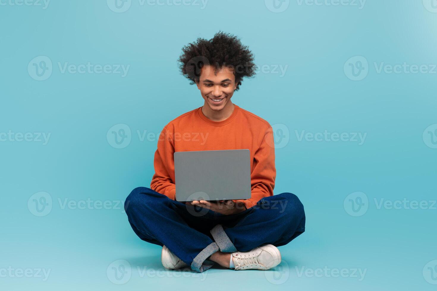 A young African American man with curly hair sits on the floor, smiling while using a laptop. He wears a casual outfit and the background is a solid light blue color. photo