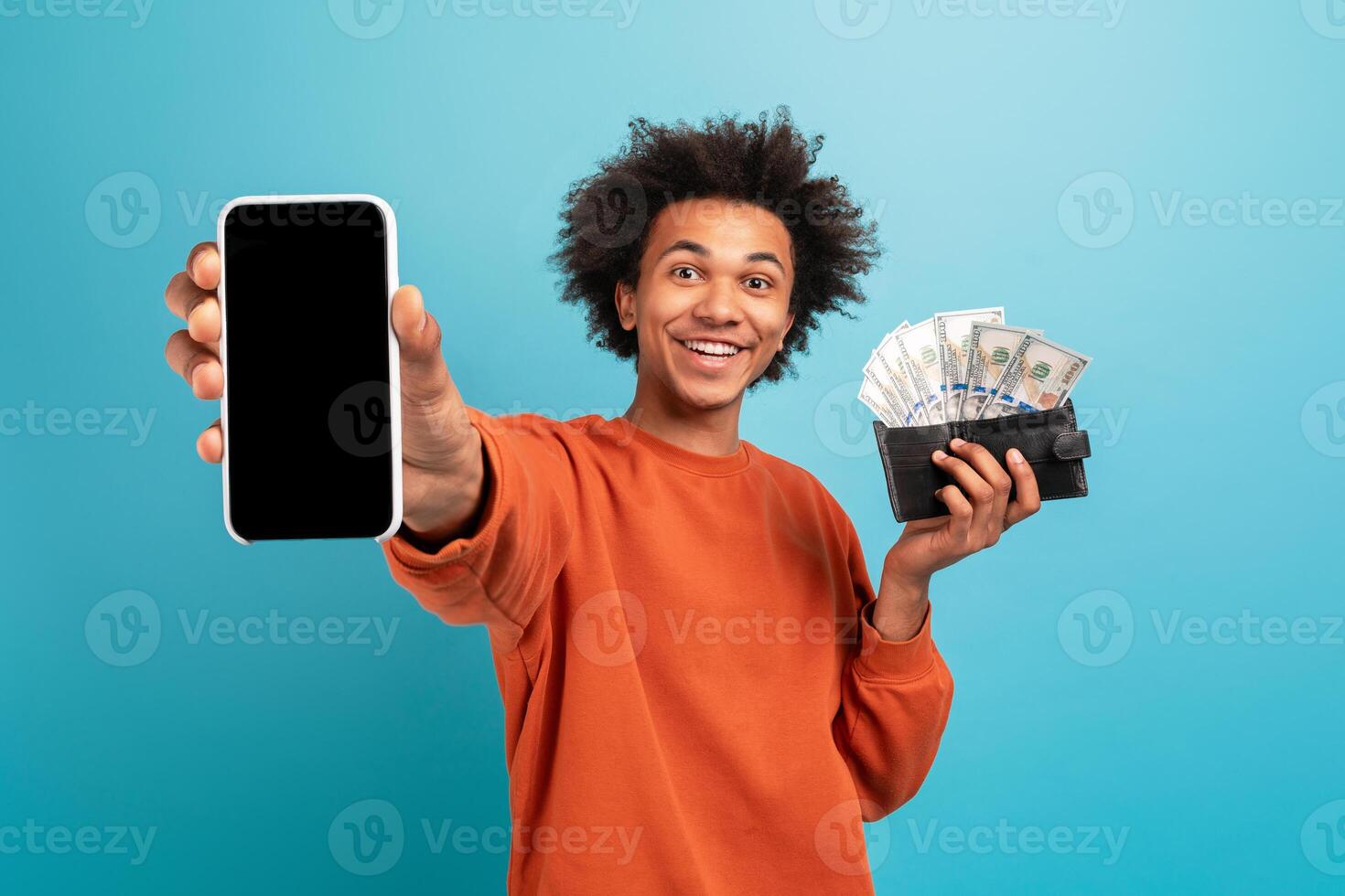 A young African American man proudly displays his smartphone in one hand and a wallet filled with cash in the other hand. He has curly hair and a bright smile, set against a blue background. photo