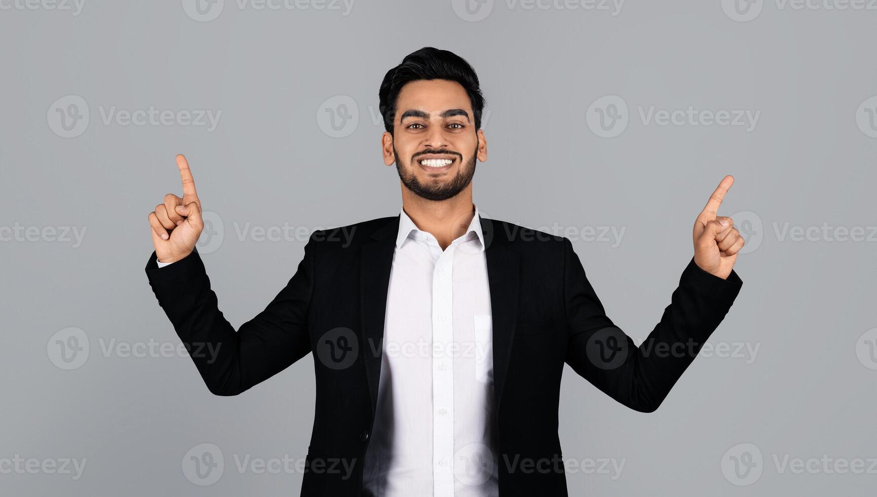 A young Arab businessman dressed in a suit stands in a studio environment, smiling and gesturing with both hands raised. His expression reflects confidence and excitement in a corporate lifestyle. photo