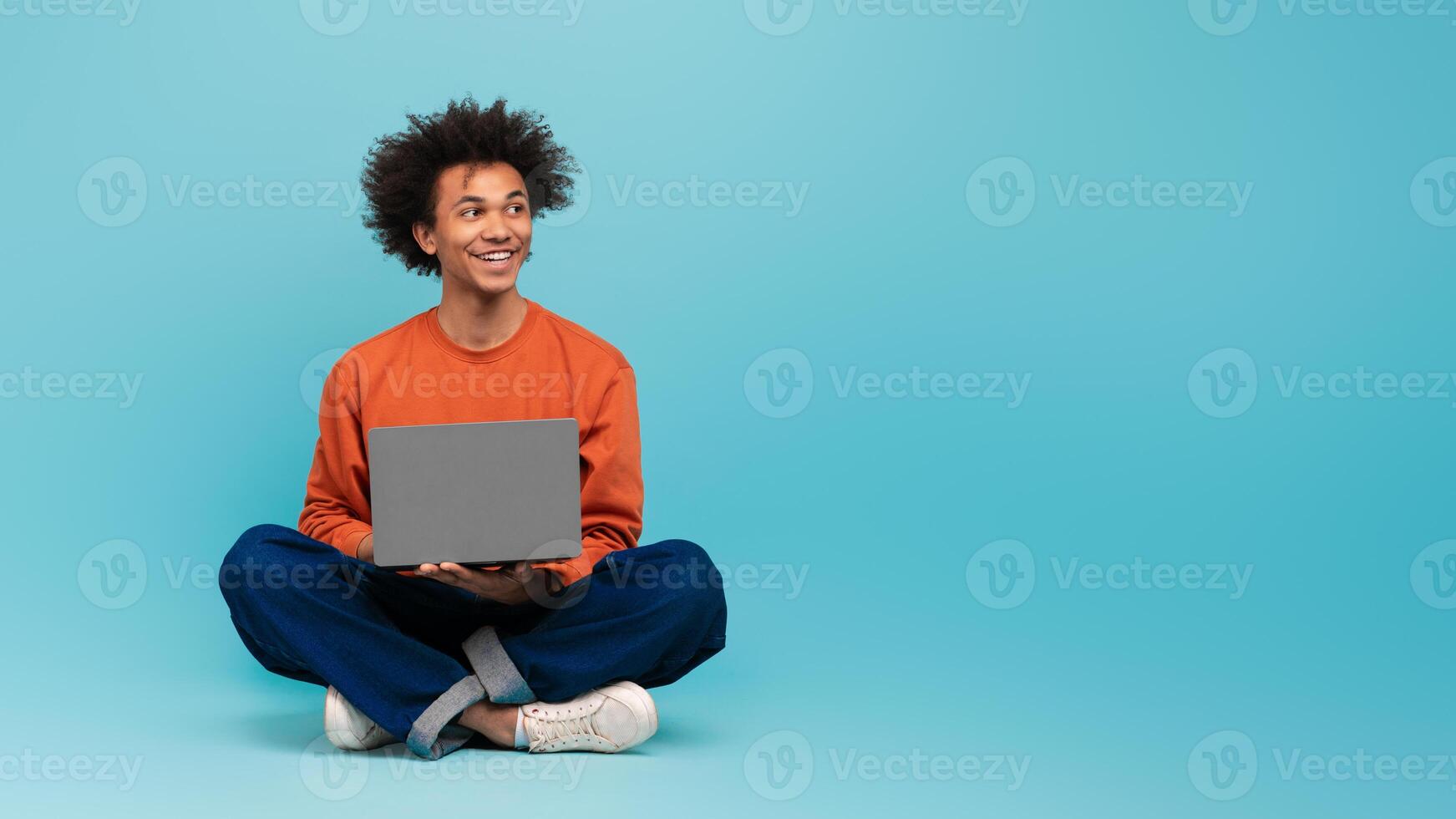 A young African American man with curly hair is seated comfortably on the floor, smiling while using a laptop against a bright blue background, copy space photo