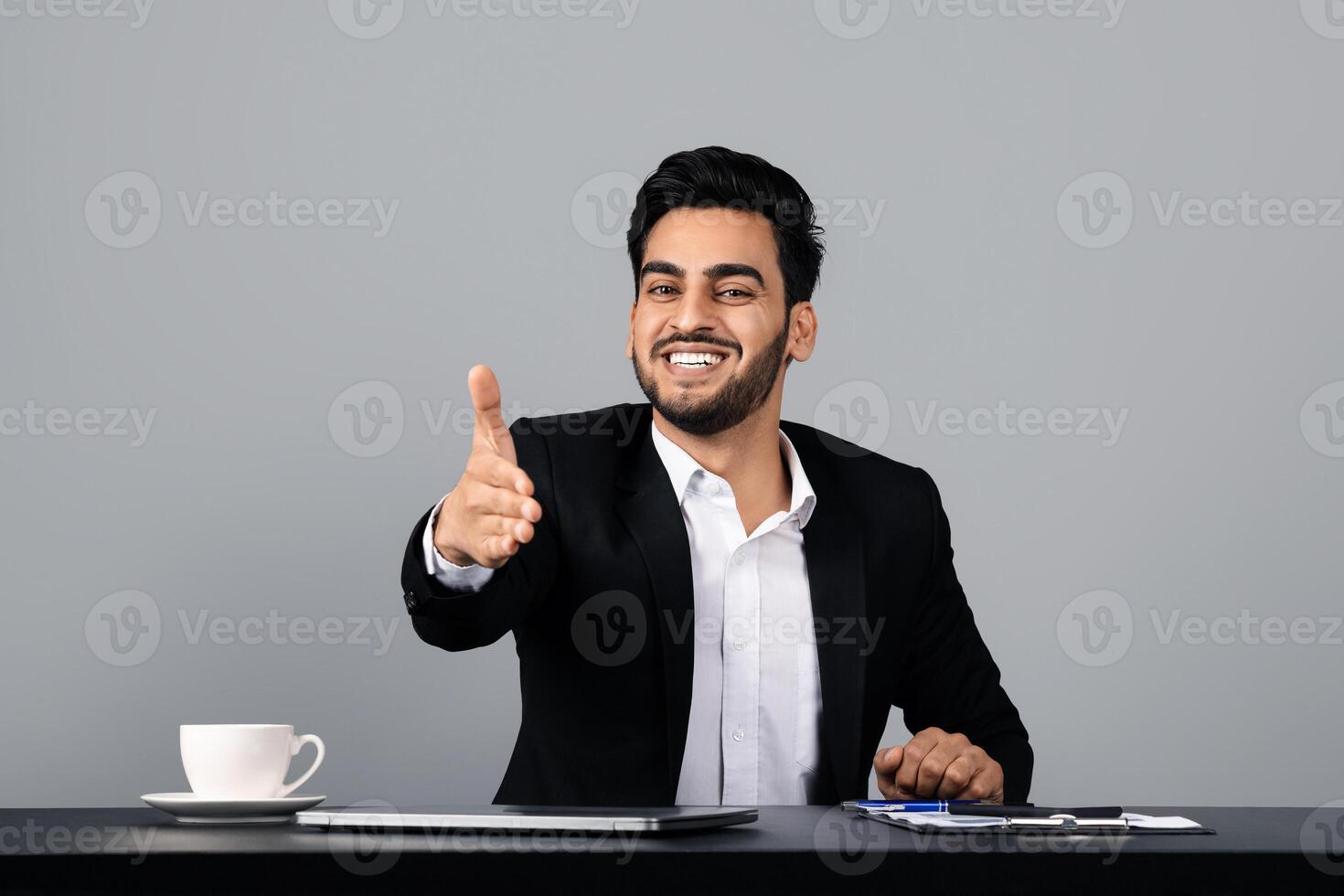 A young Middle Eastern man dressed in a suit extends his hand for a handshake, smiling warmly. He sits at a desk in a modern studio, ready for professional engagement. photo