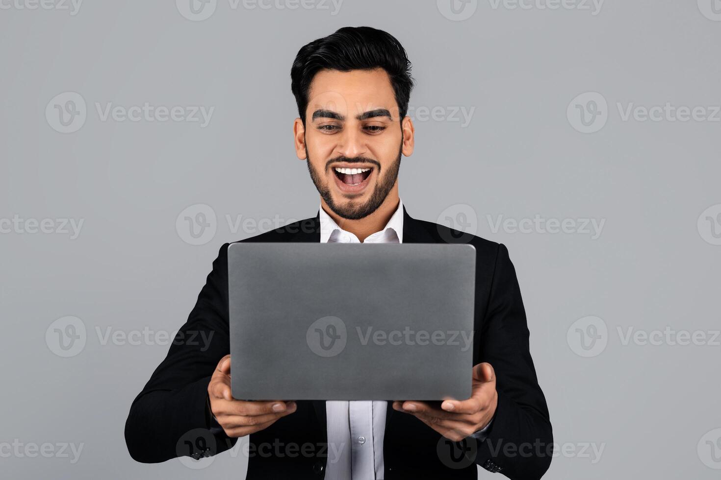 A Middle Eastern businessman smiles broadly while working on his laptop in a minimalistic studio. Dressed in formal attire, he shows excitement over a recent achievement. photo