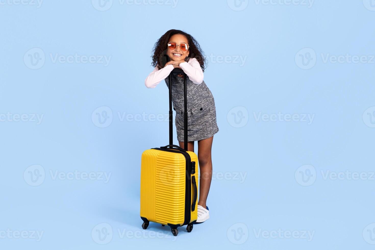 A joyful African American girl stands with a yellow suitcase, leaning on it with a big smile. She wears stylish sunglasses and is dressed in a playful outfit, exuding excitement for her adventure. photo