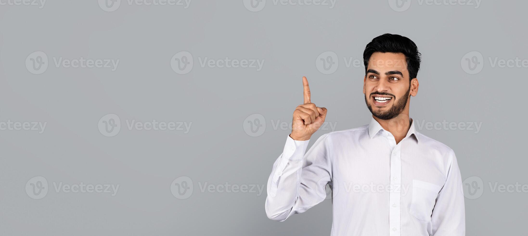 A Middle Eastern businessman in a white shirt smiles while standing in a studio. He confidently points at copy space, suggesting an idea or innovation in his corporate environment. photo