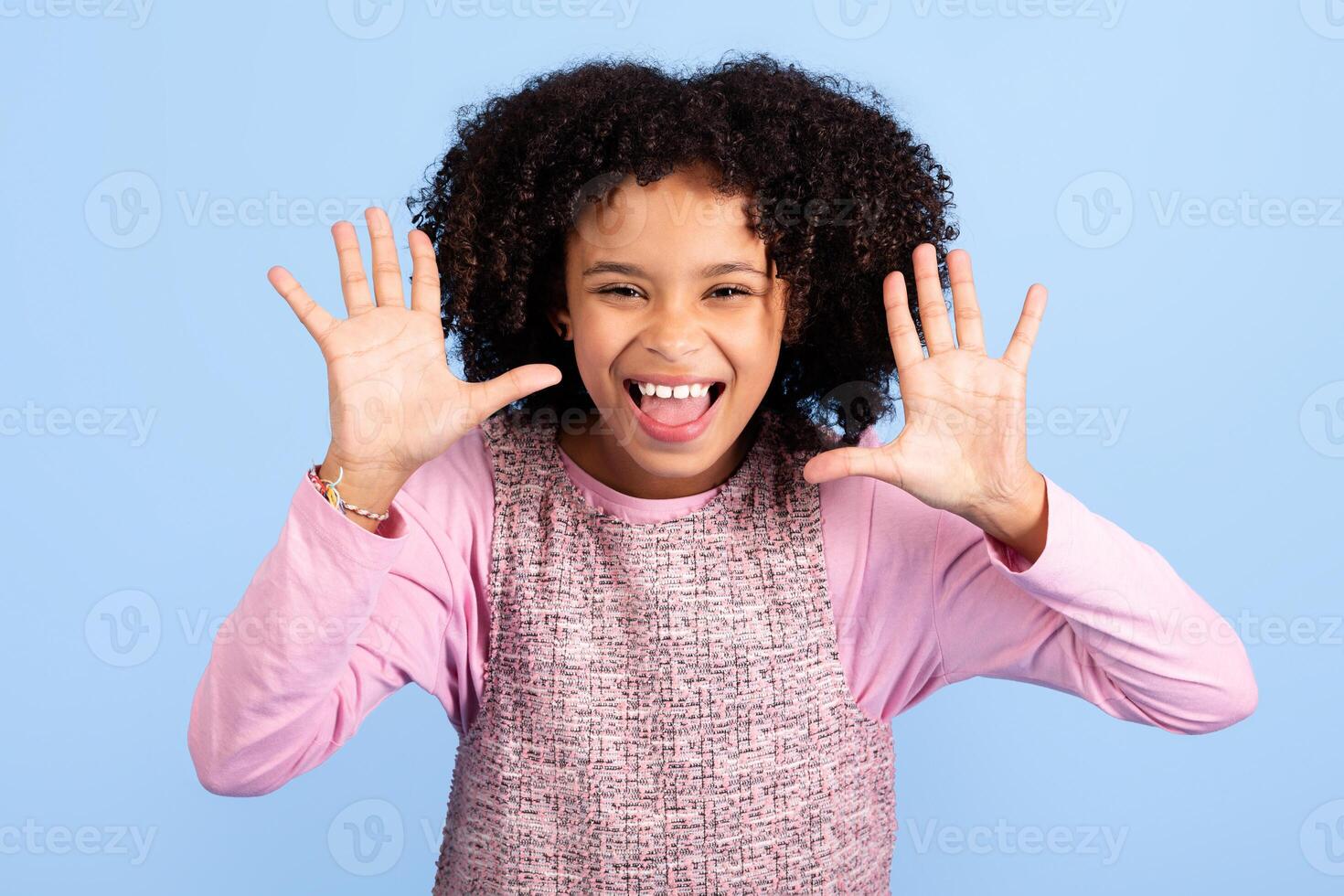 A joyful girl with curly hair displays her hands enthusiastically, expressing fun and happiness. She is dressed in a pink top and stands in front of a solid blue backdrop, radiating positivity. photo