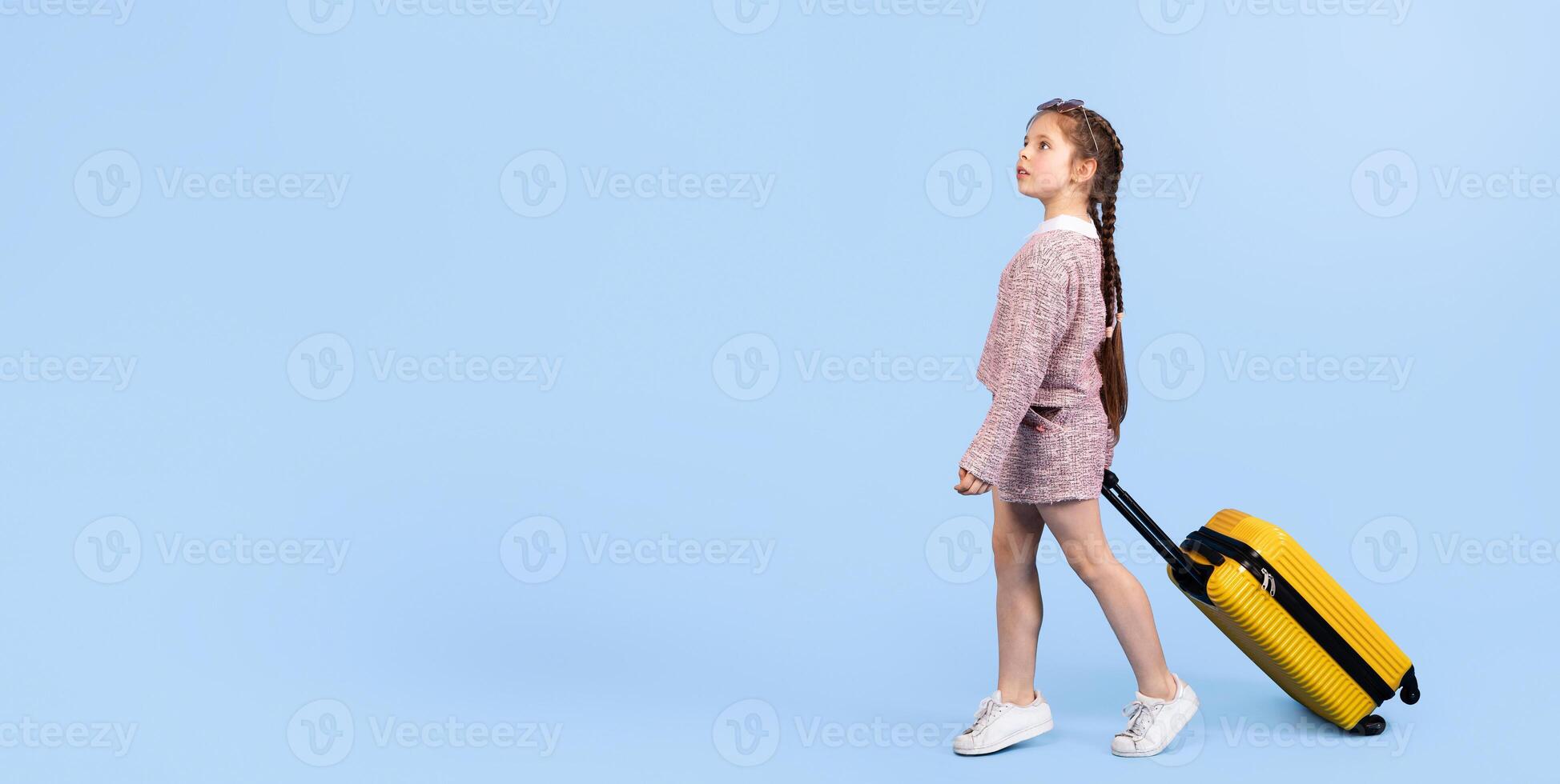 A joyful preteen girl with braided hair is pulling a yellow suitcase against a light blue backdrop. She radiates happiness, embodying the carefree spirit of childhood and adventure, copy space photo