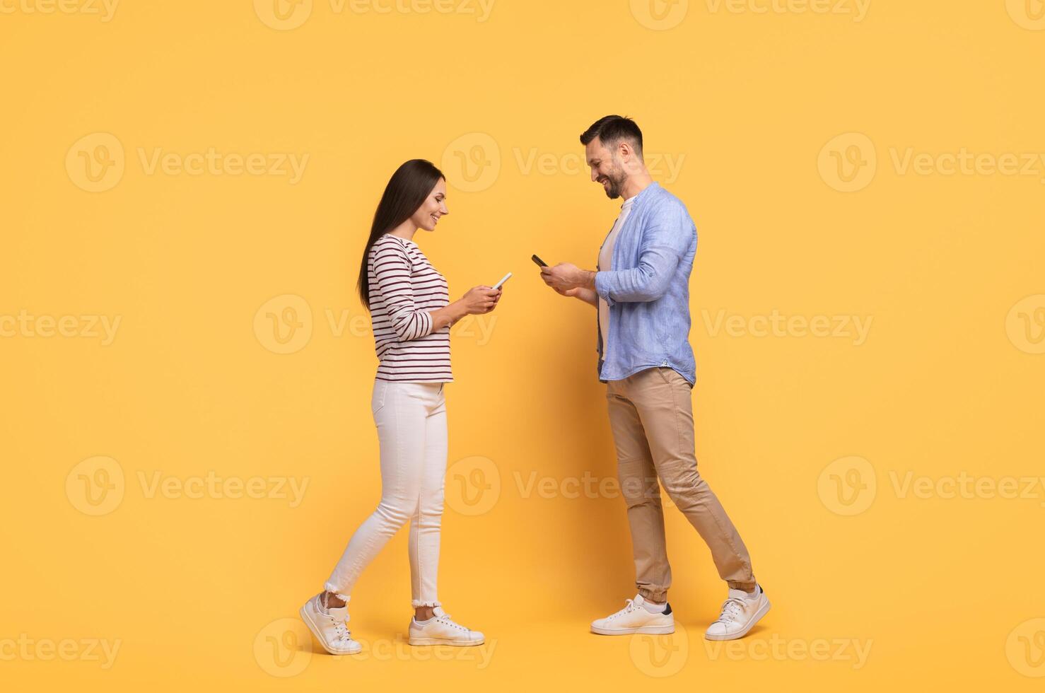 A family couple stands close together against a vibrant yellow backdrop, engaging with their smartphones. They share smiles and laughter, reflecting their affectionate bond and enjoyment. photo
