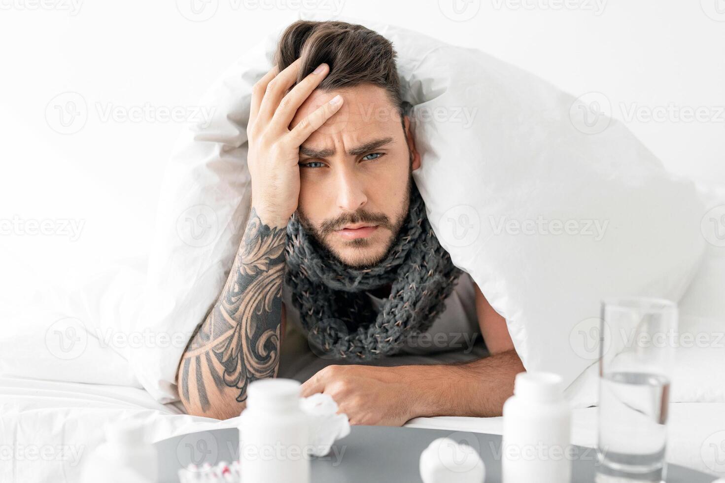 Sick young man feeling unwell, touching forehead and looking at camera, lying in bed under blanket, table full of pills. Covid-19 coronavirus and seasonal flu symptoms concept photo