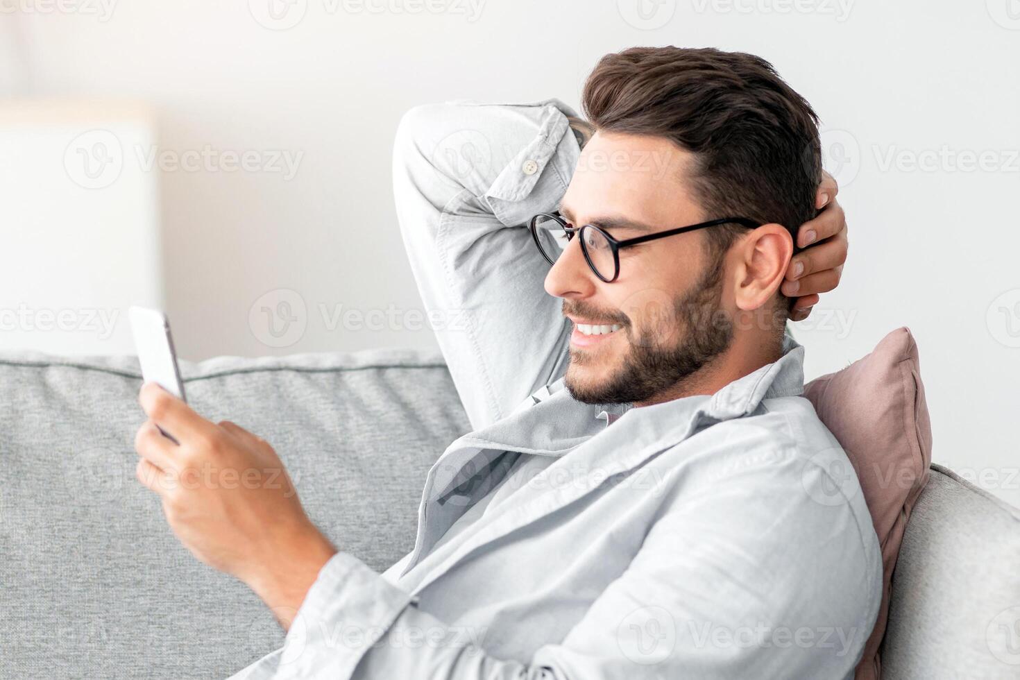 Cool gadget and app. Young smiling guy holding mobile phone, typing sms message, resting on sofa in living room, free space. Man browsing internet, surfing web, using app photo