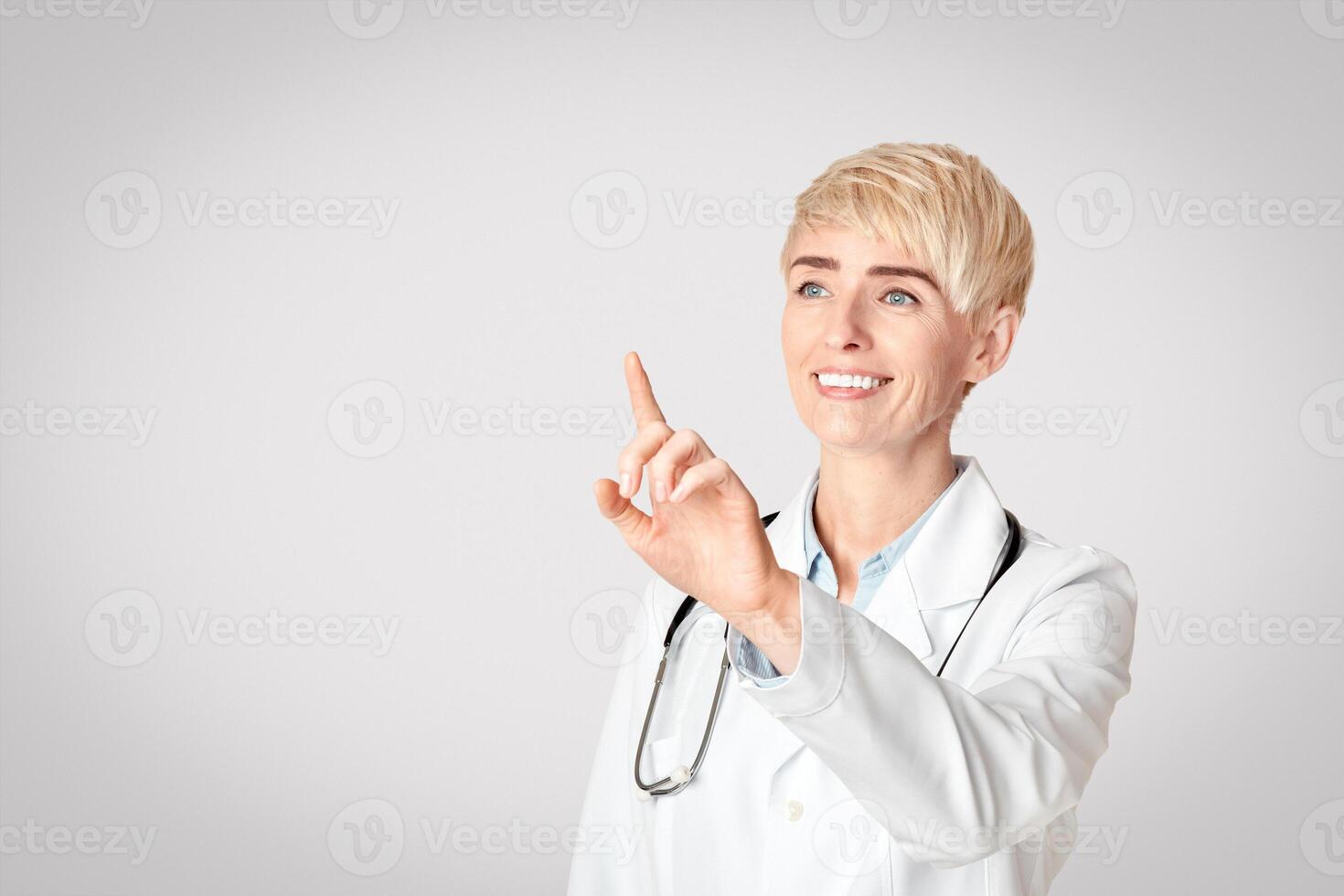 Modern medicine, diagnostics and science. Smiling adult female doctor in coat raises her hand and press fingers an empty space for virtual button, or board, or invisible screen, studio shot photo