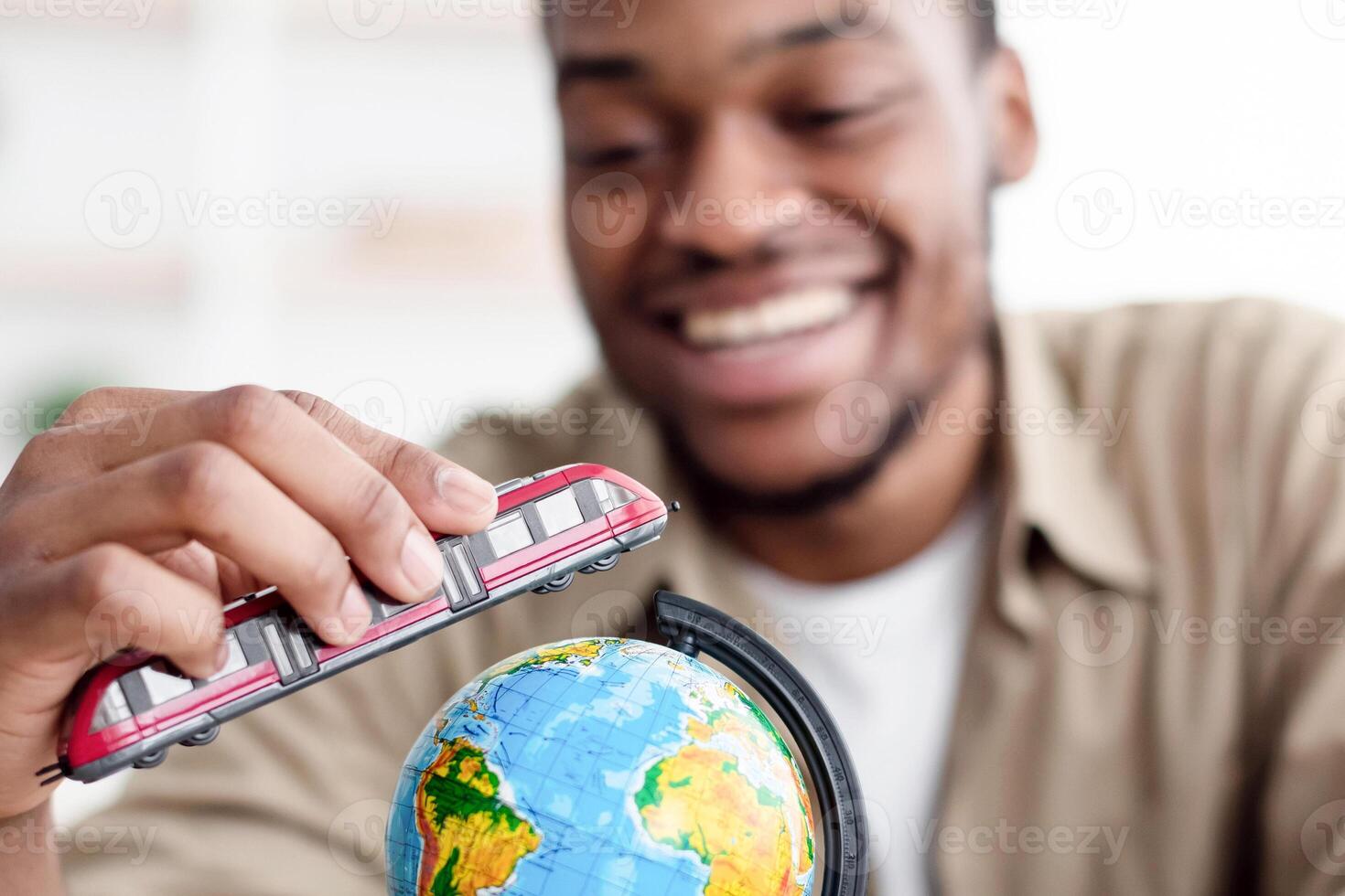 Travel. Cheerful African Guy Playing With Train Model And Globe Sitting At Desk Indoors, Planning His Next Trip. Black Man Dreaming About Journey And Vacation Concept. Selective Focus On Toy photo