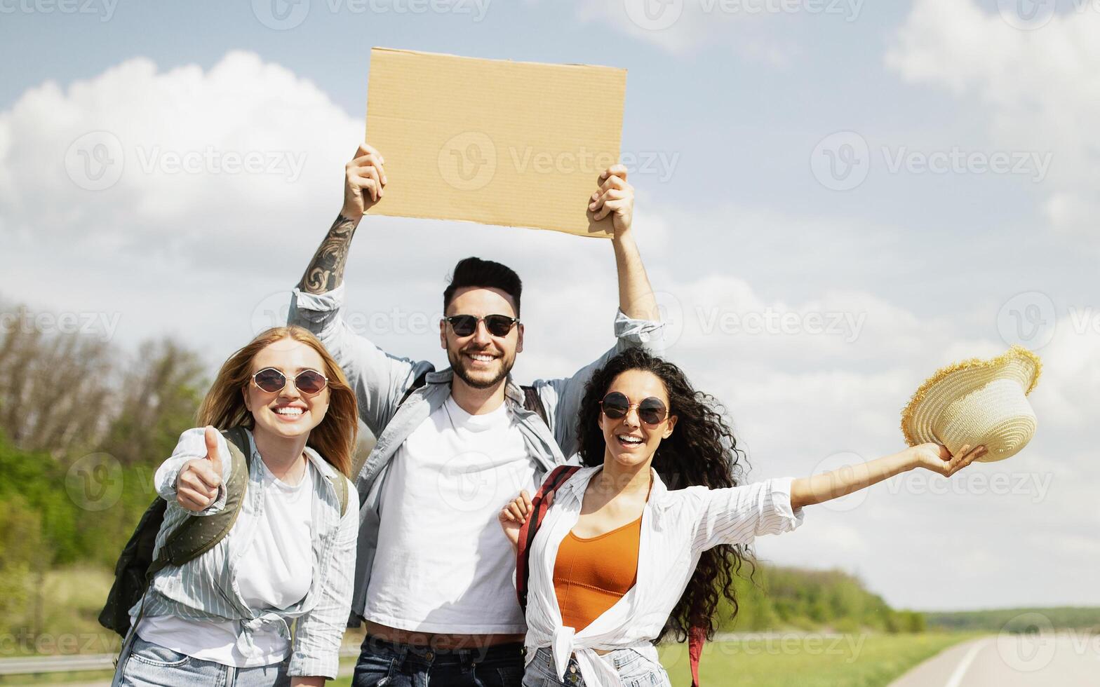 Joyful multiethnic friends thumbing their ride on highway with empty sign, playing guitar, having fun, going on autostop journey together, outdoors. Group of hitchhikers having fun on road trip photo