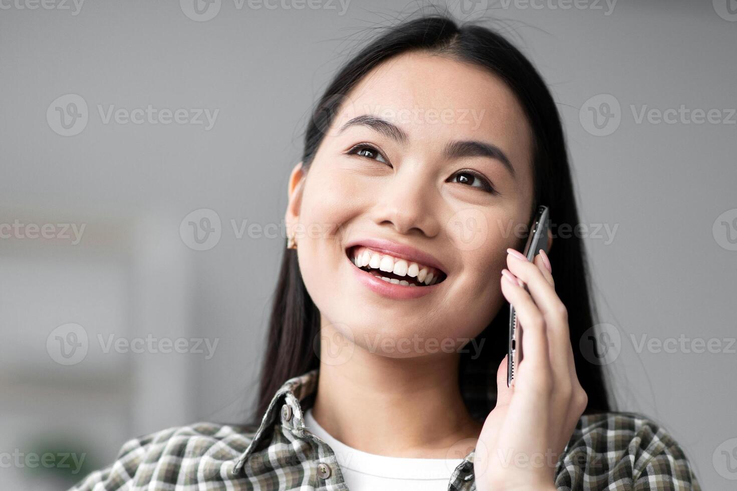 Happy asian young woman sitting on couch and having phone conversation, looking at copy space. Cheerful korean lady talking on phone while relaxing on sofa in living room, closeup portrait photo
