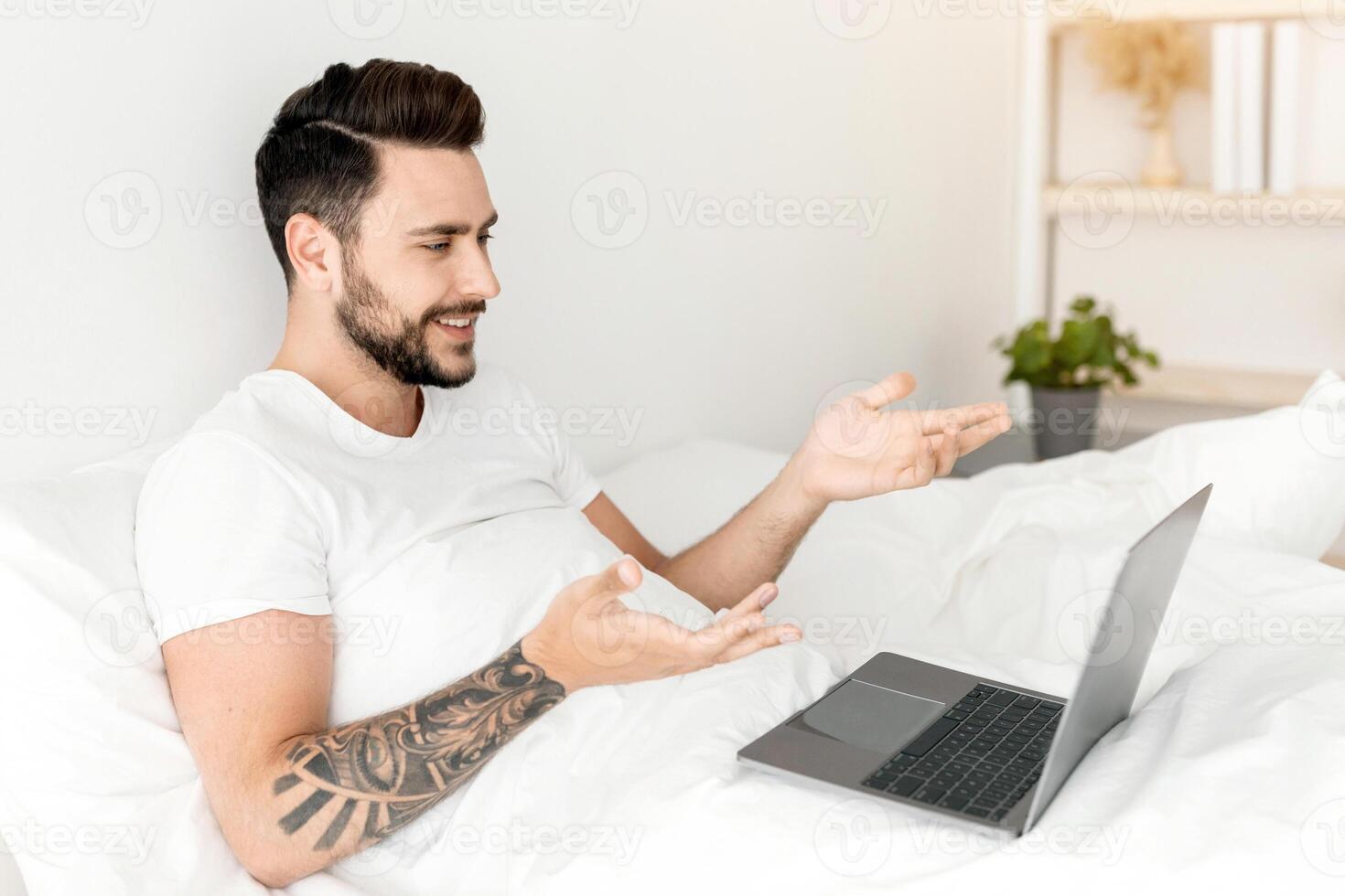 Young man having call on laptop computer while sitting in bed at home on weekend morning and talking to family distantly. Modern communication concept photo