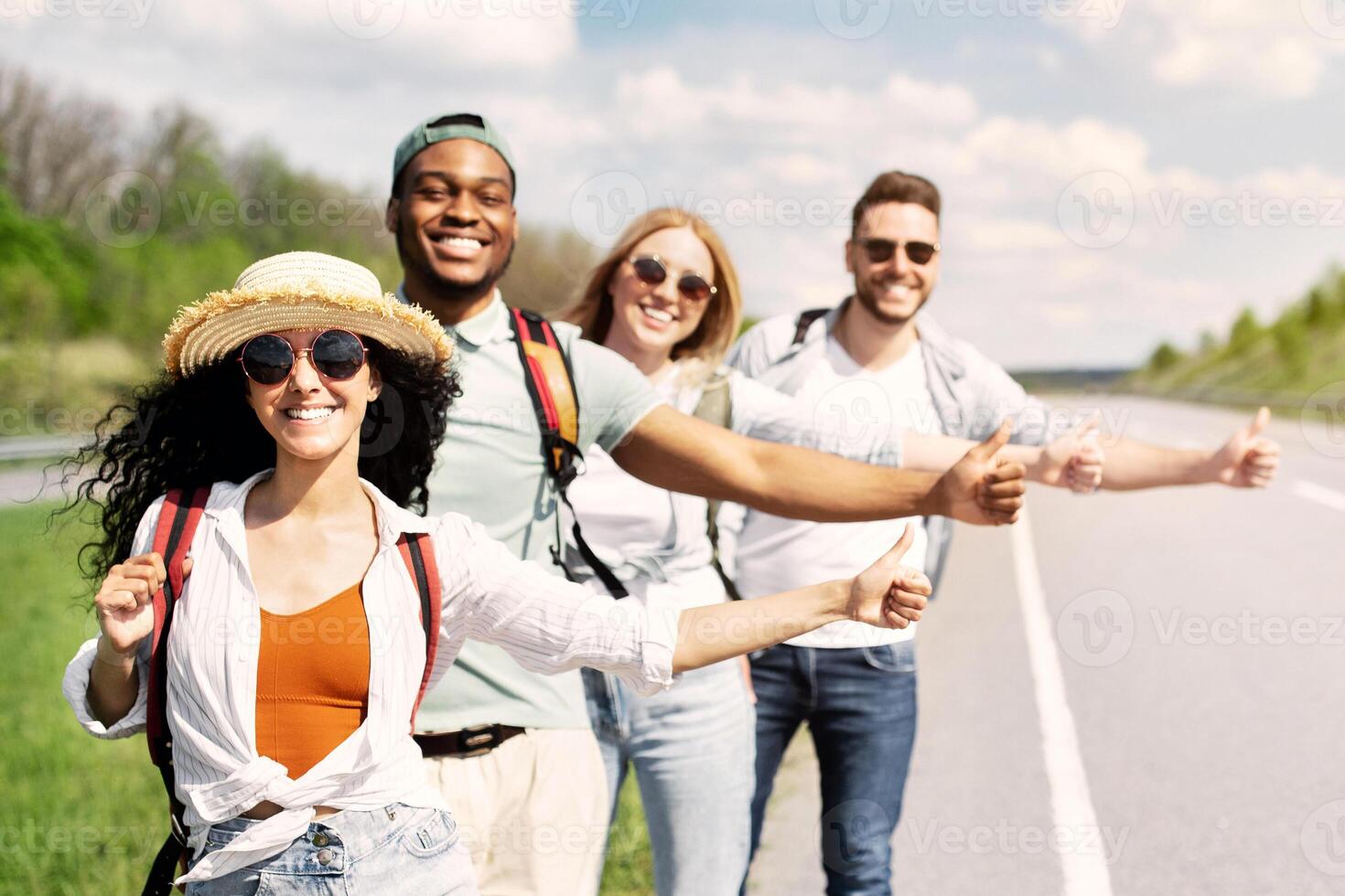 Young multiracial friends standing on highway, showing hitchhiking gesture, stopping car, going on journey together, copy space. Traveling by autostop, summer vacation, tourism and friendship concept photo