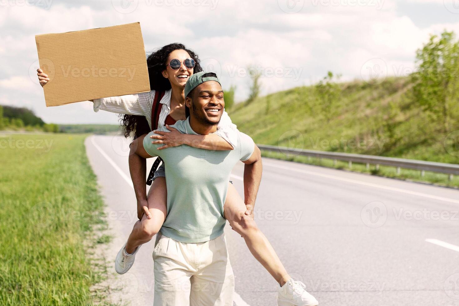 Millennial multiracial couple traveling by autostop, hitchhiking, looking for transport, trying to stop car, holding empty cardboard sign with mockup, ready strat new journey, copy space photo