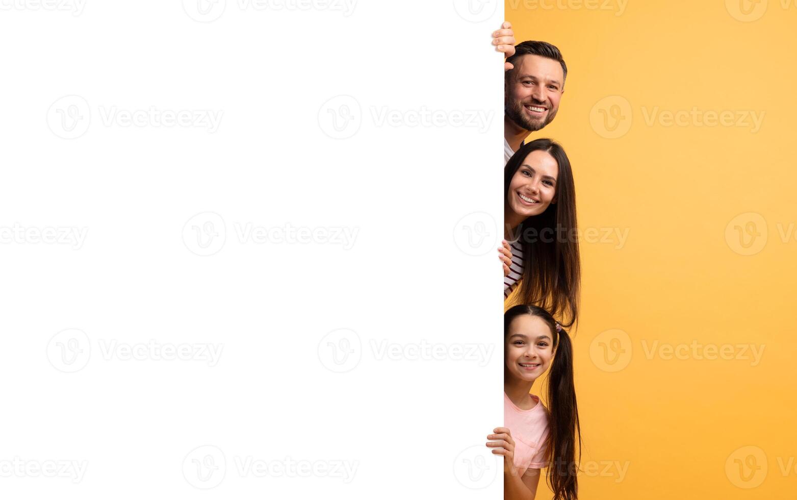 A cheerful family of four stands together, playfully peeking around a large blank surface. Their smiles show affection and happiness in a vibrant yellow background, creating a joyful atmosphere. photo
