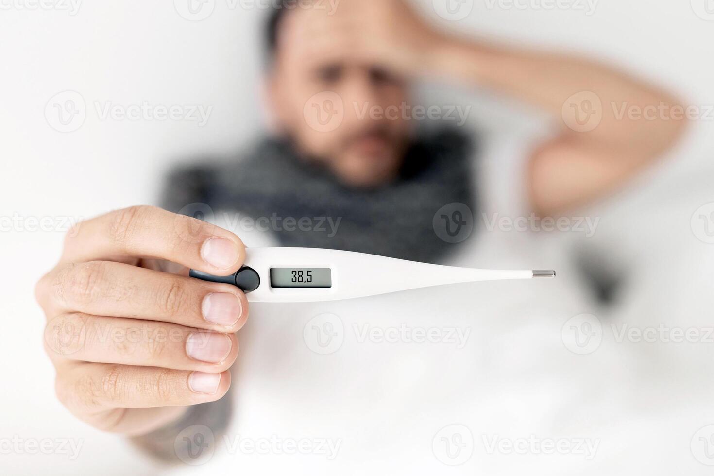 Sick young man showing thermometer to camera, lying on bed and touching forehead. Coronavirus, influenza symptom concept. Selective focus, top view photo
