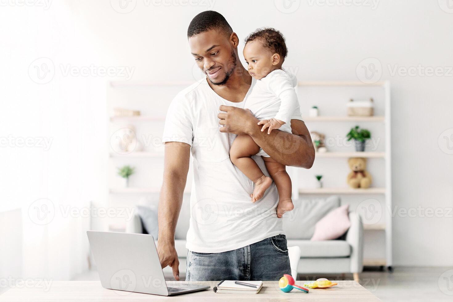 Young Black Man Holding Little Baby On Hands And Using Laptop At Home, Millennial African American Father Working Remotely With Computer While Babysitting His Adorable Infant Child, Copy Space photo