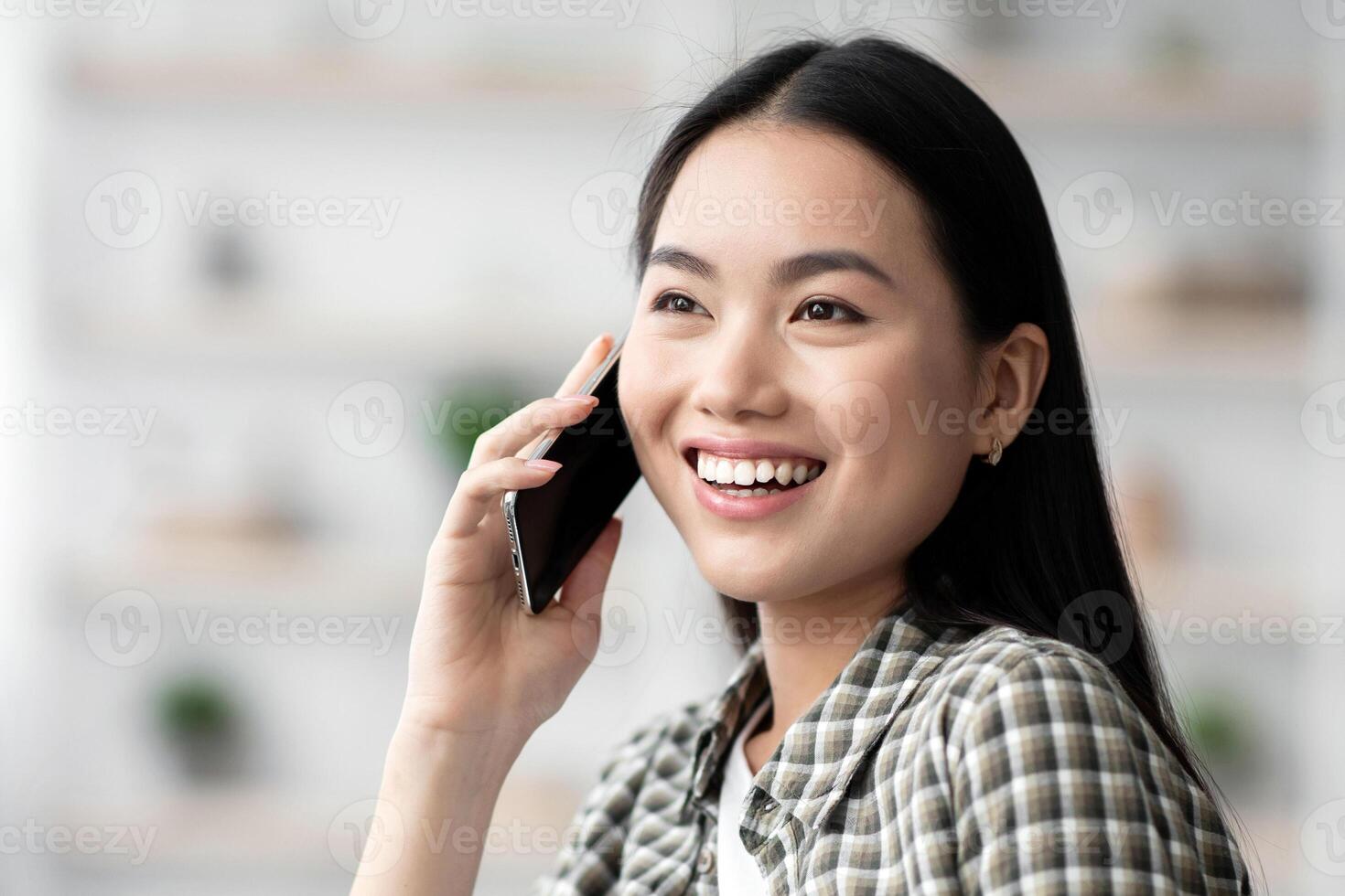 Pretty young asian woman chilling on couch at home, talking on cellphone, smiling and looking at copy space, closeup portrait. Happy chinese lady having phone conversation with girlfriend or lover photo
