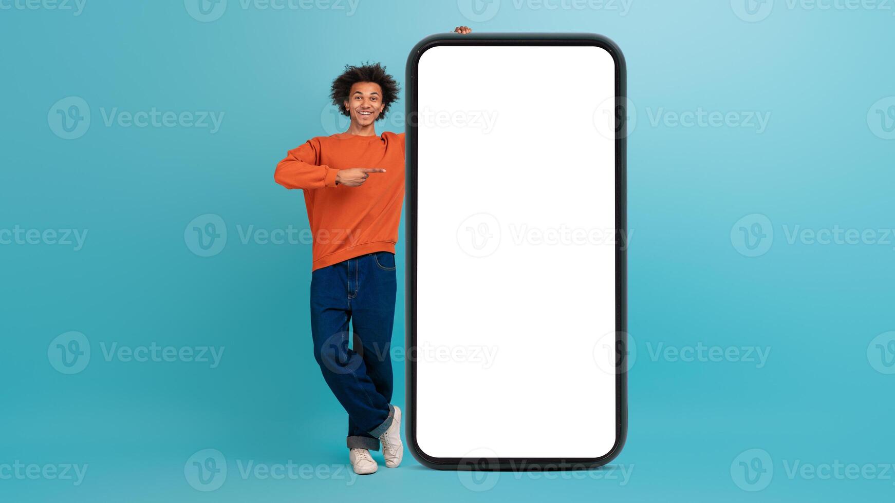 A young African American man with curly hair stands beside a large blank smartphone display. photo