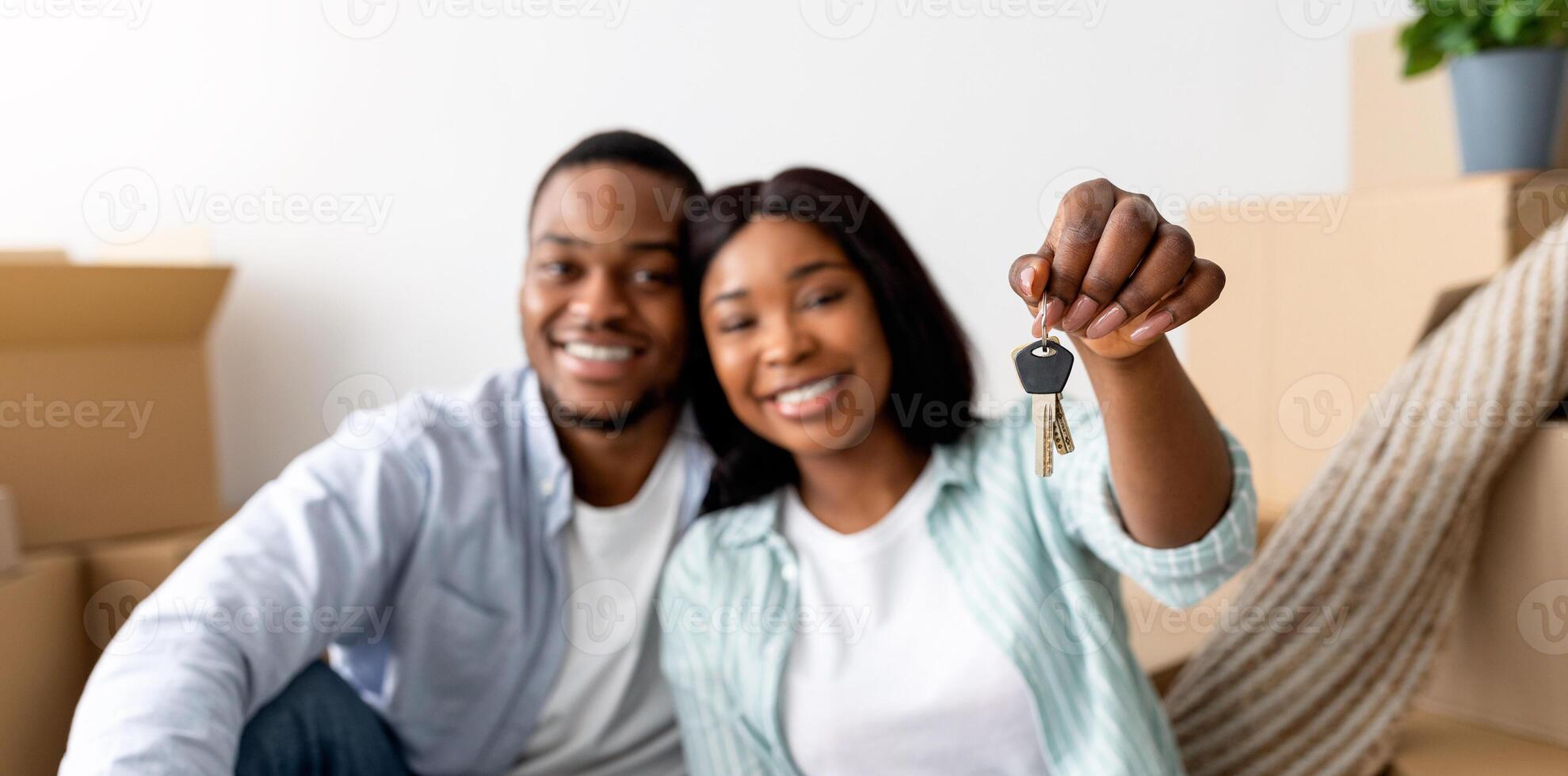 Excited black married couple showing home keys while sitting among cardboard boxes, selective focus. Happy spouses celebrating moving to new apartment, enjoying relocation photo