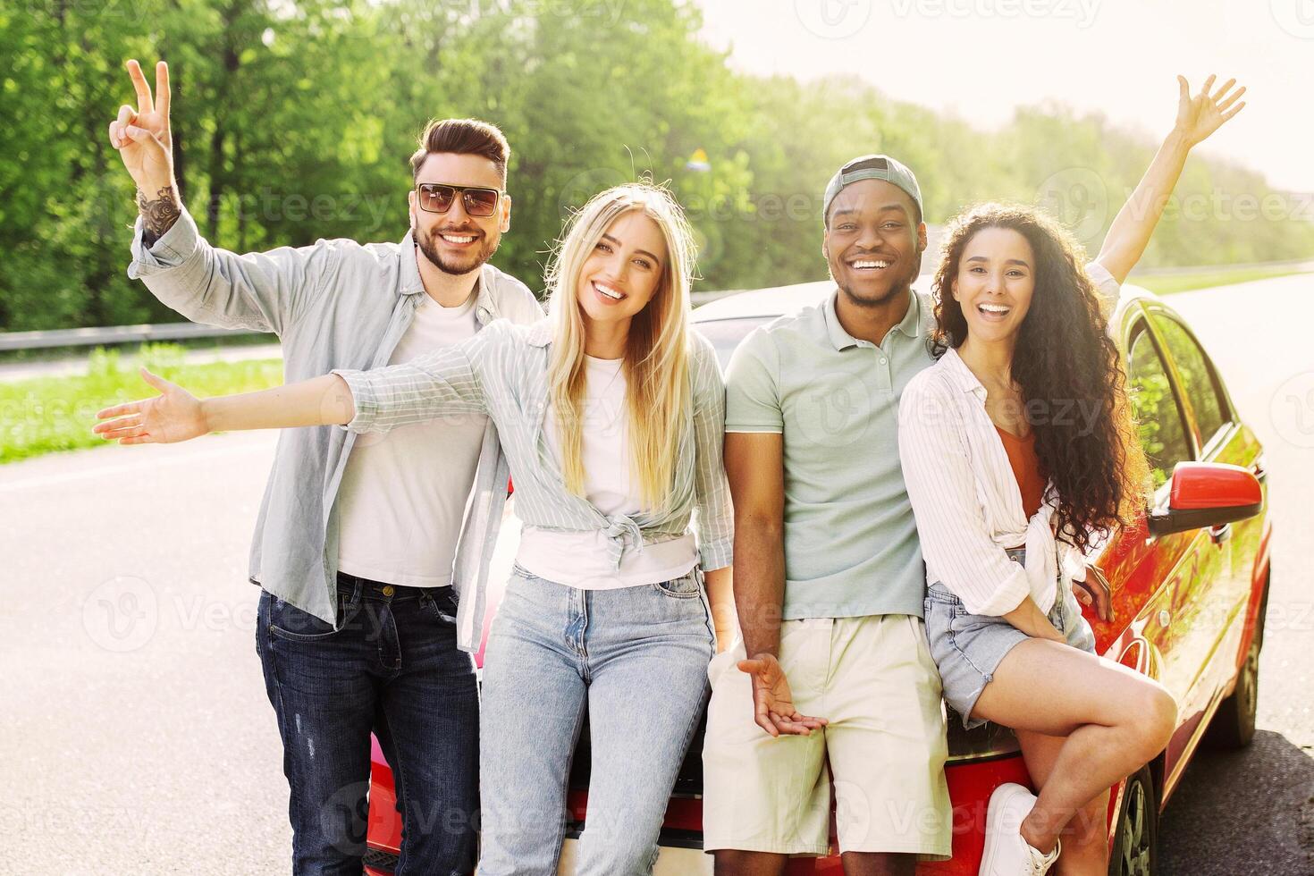 Portrait of cheerful multiethnic friends posing near their car on roadside, smiling at camera, panorama. Adventurous young students having road trip in summertime, hitchhiking together photo