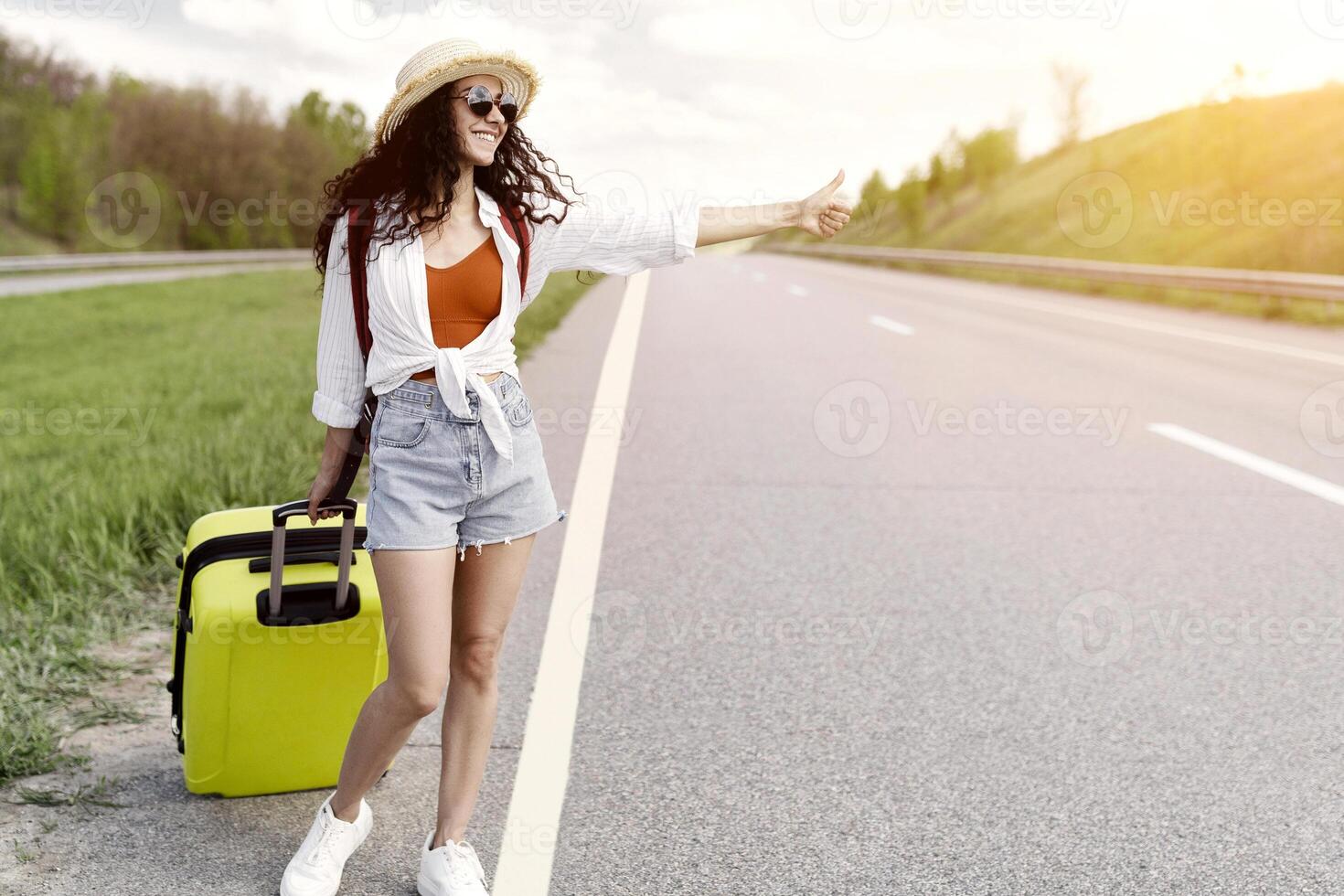 Full length of happy young woman with backpack and suitcase trying to stop car, showing thumb up, asking for free ride, hitchhiking on roadside, copy space. Traveling by autostop concept photo