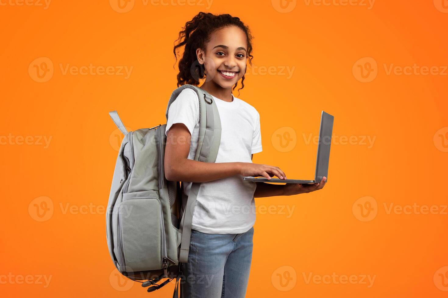 E-learning Concept. Portrait of smiling African American teenage girl holding and using laptop over yellow orange studio background, looking at camera, typing on keyboard, wearing backpack photo