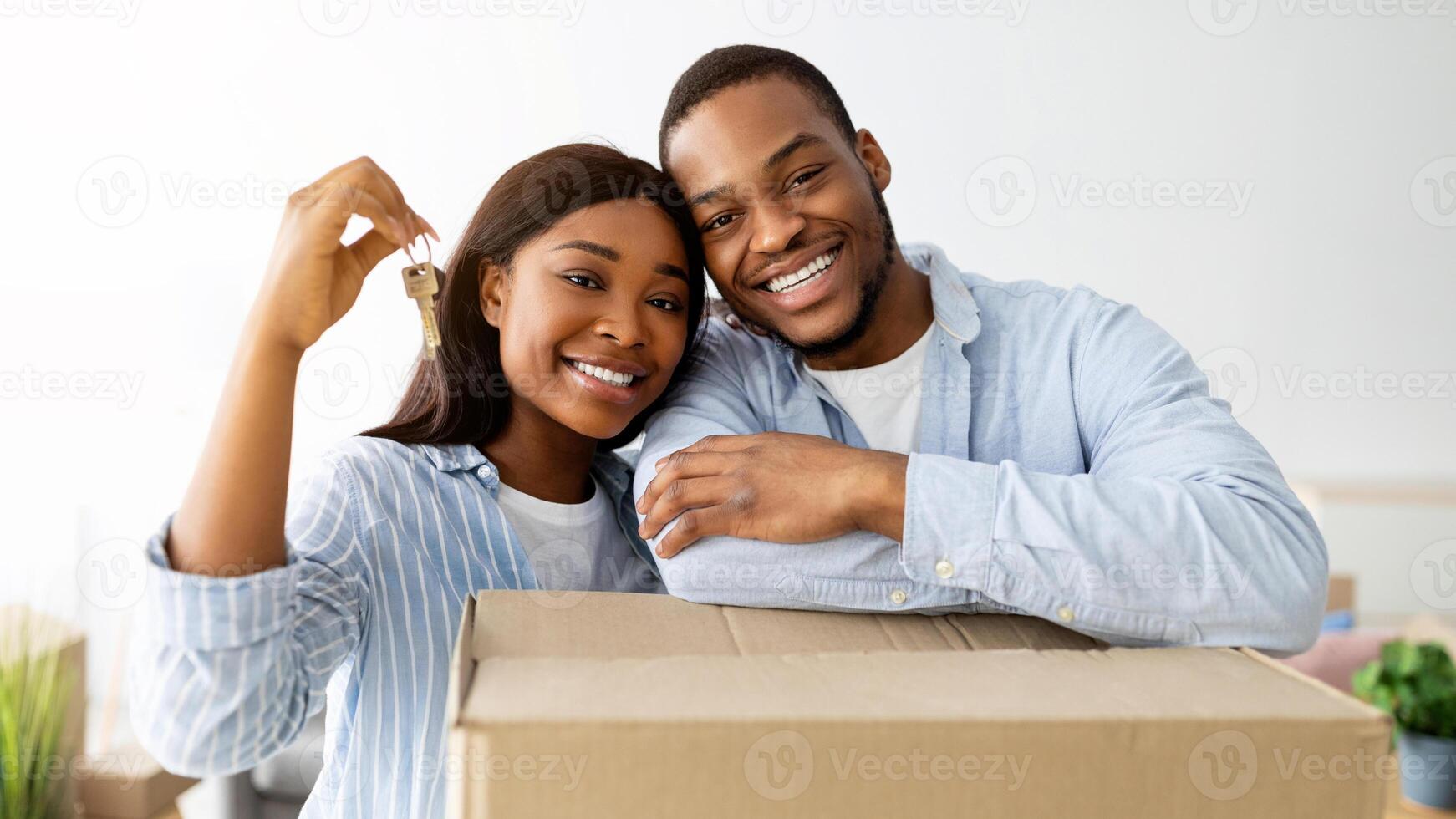 Real estate concept. Loving african american young spouses posing with moving boxes and new house key, hugging and smiling to camera, standing in room full of boxes photo