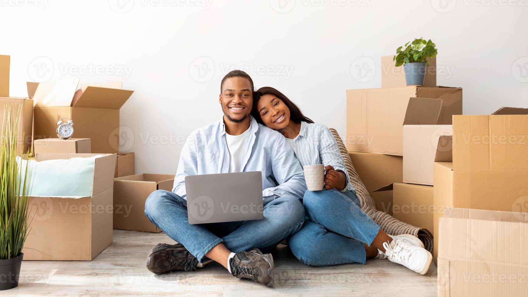 Moving concept. Happy african american couple purchasing furniture for new apartment, sitting on floor and using laptop computer, free space. Excited spouses smiling to camera, sitting among boxes photo