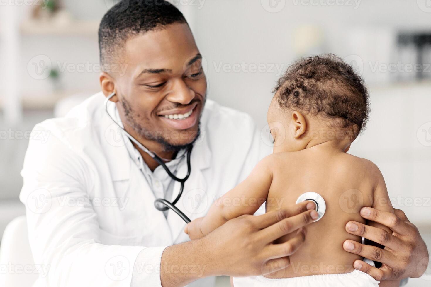 Portrait Of Handsome Black Pediatrician Examining Baby Patient With Stethoscope, Friendly African American Doctor Checking Little Infant Child's Health During Check Up In Clinic, Closeup Shot photo