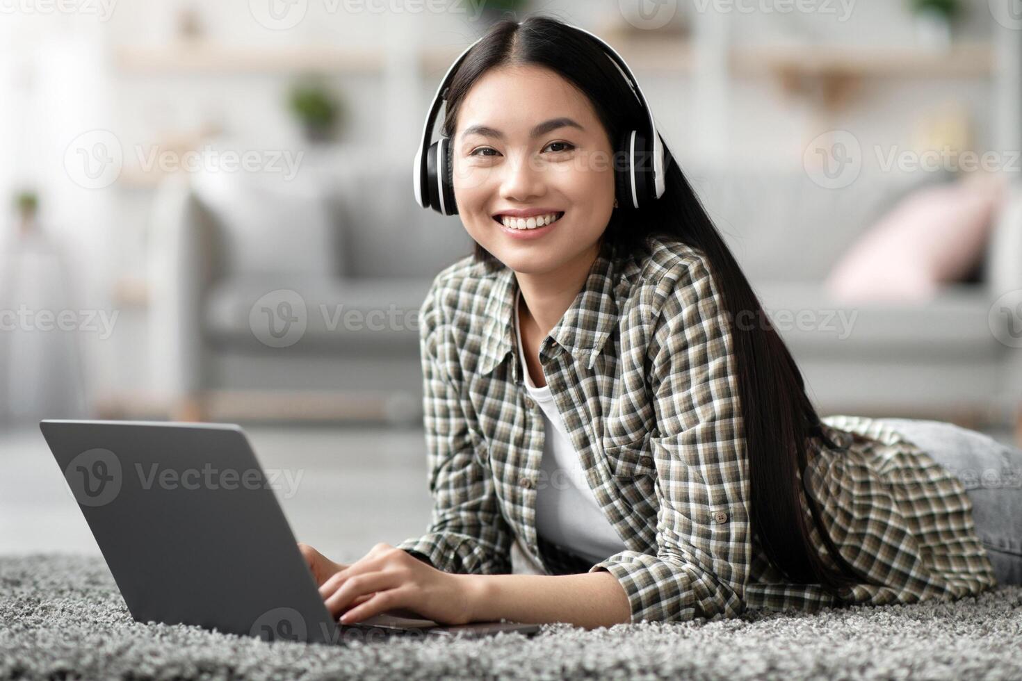 Relaxed asian lady using wireless headset and laptop while chilling at home alone, laying on carpet in living room, playing games or watching movie online, copy space. Domestic entertainment photo