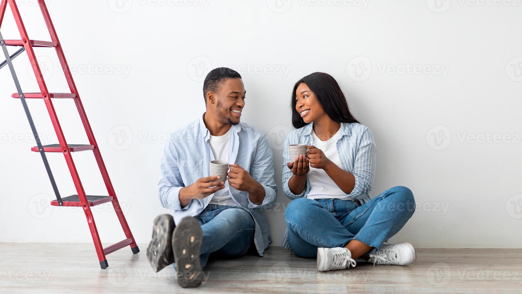 Young african american couple discussing plan of repairs in their new house and drinking coffee while sitting on the floor near ladder. Loving spouses having break after doing renovation in new flat photo