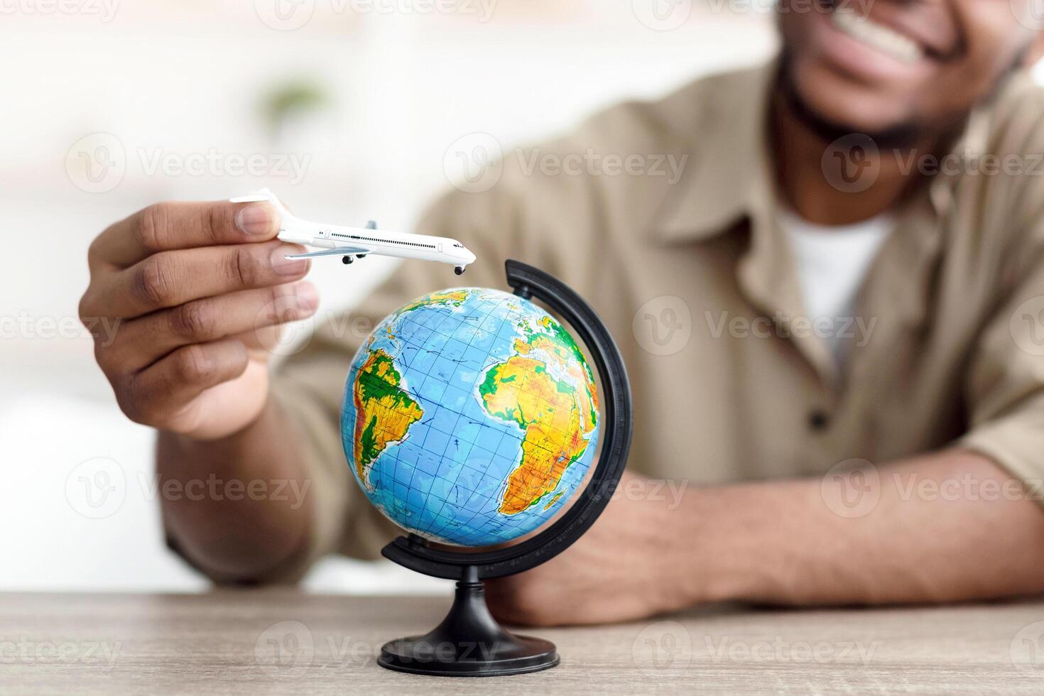 Cheerful Black Traveler Guy Playing With Airplane Toy Model And World Globe Sitting At Desk At Home. Vacation Planning, Touristic Destinations Concept. Selective Focus, Shallow Depth photo