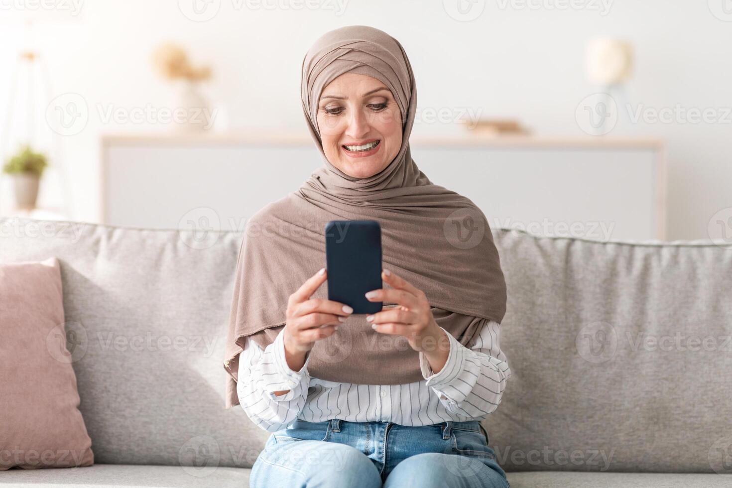 Happy Senior Muslim Woman Using Phone Texting And Browsing Internet Sitting On Couch At Home, Wearing Traditional Hijab. New Mobile Application For Smartphone Advertisement Concept photo