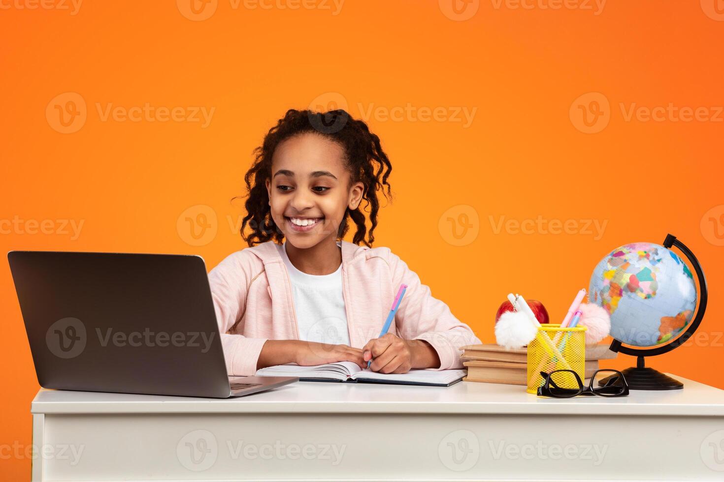 Remote Learning. Portrait of positive African American schoolgirl sitting at desk, writing in notebook and using laptop computer. Smiling young black girl watchin online course or having virtual class photo