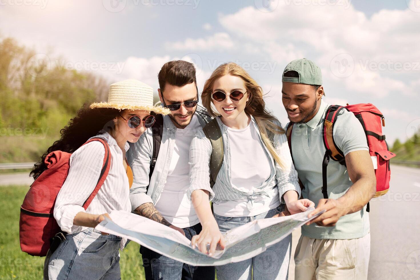 Group of best friends with backpacks checking map during road trip, hitchhiking on highway. Multiracial young tourists plotting their route, having autostop journey on summer holidays photo