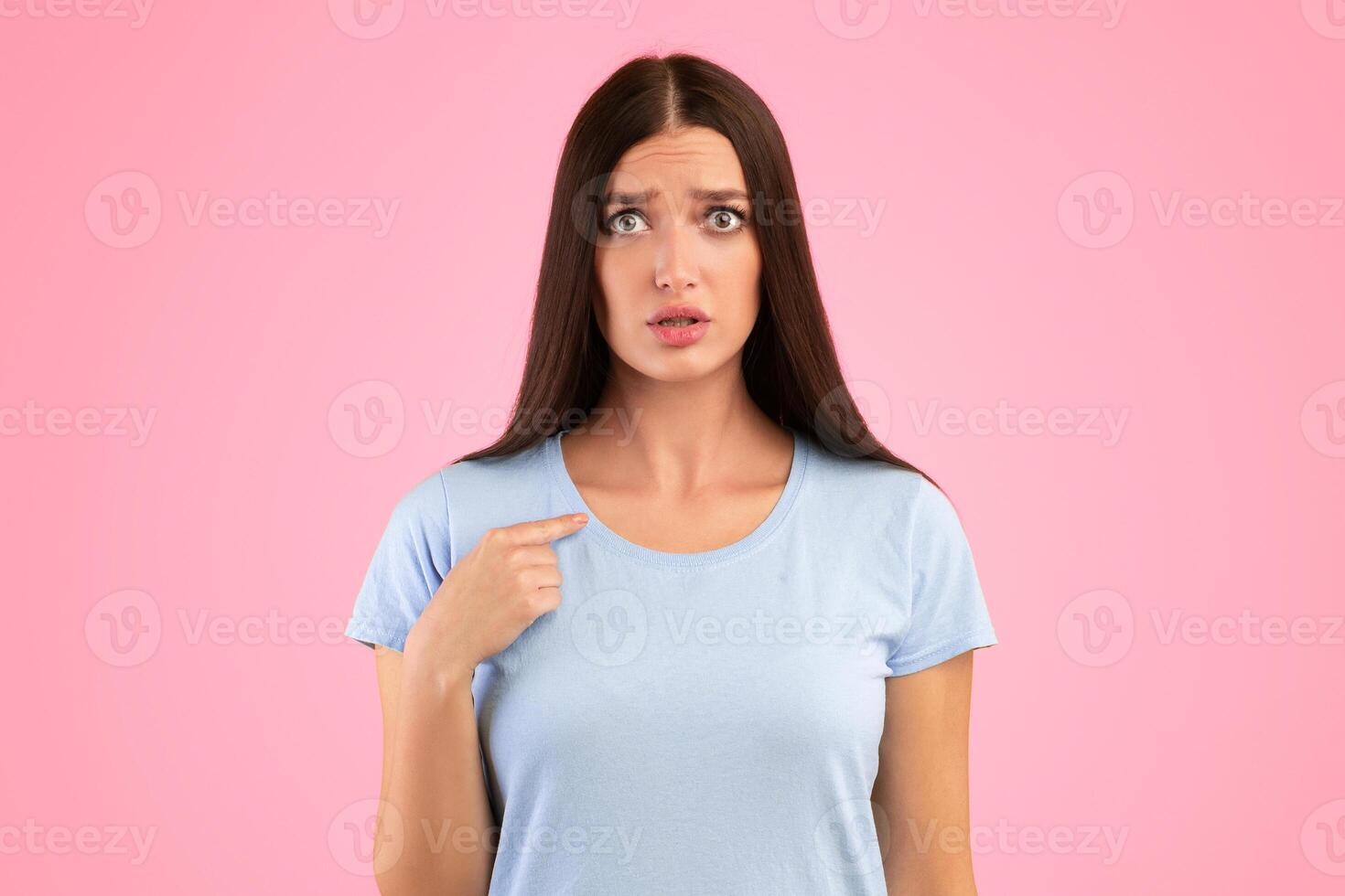 Are You Looking At Me. Portrait of confused young lady pointing finger at herself, posing at camera, standing isolated over orange studio background. Surprised scared female model indicating her photo