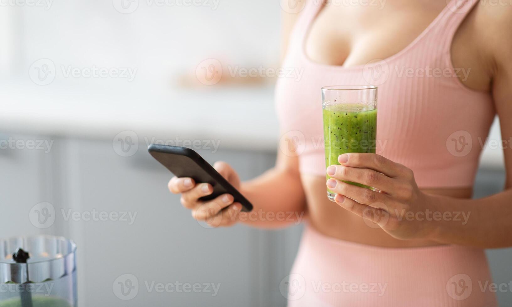 A woman in a pink top holds a glass of green smoothie in one hand and a smartphone in the other. She appears to be enjoying her drink while possibly browsing content in her kitchen, cropped photo