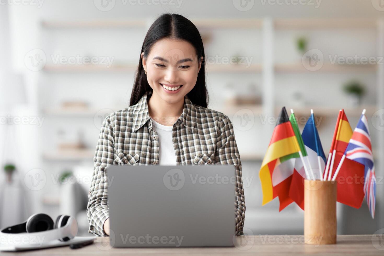 Positive asian young woman foreign language teacher sitting at workdesk with international flags and using laptop at home, smiling at screen, copy space. Foreign language online course, class concept photo