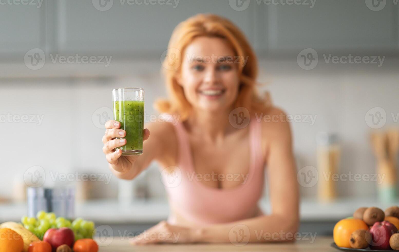A cheerful woman holds a glass of green smoothie in a bright kitchen. Various fresh fruits are arranged on the countertop, showcasing a healthy lifestyle and vibrant food choices. photo