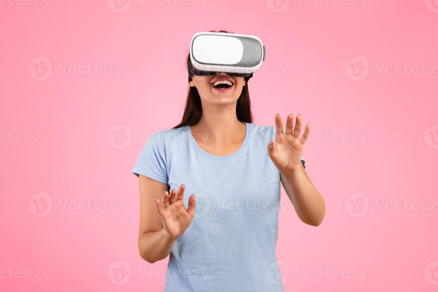 Excited young woman in VR headset touching air during virtual reality experience at studio. Emotional lady exploring artificial computer world or playing game, isolated on yellow background photo