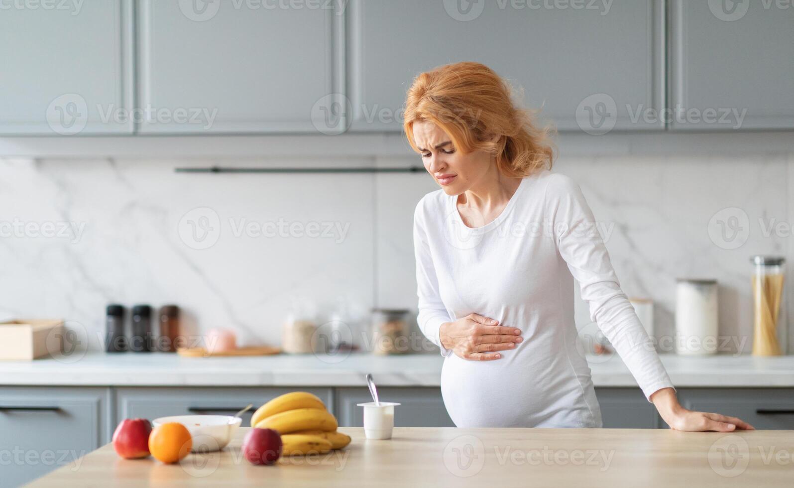 A pregnant woman is standing in a modern kitchen, visibly uncomfortable as she holds her belly while looking down at a kitchen counter filled with fruit and yogurt, copy space photo