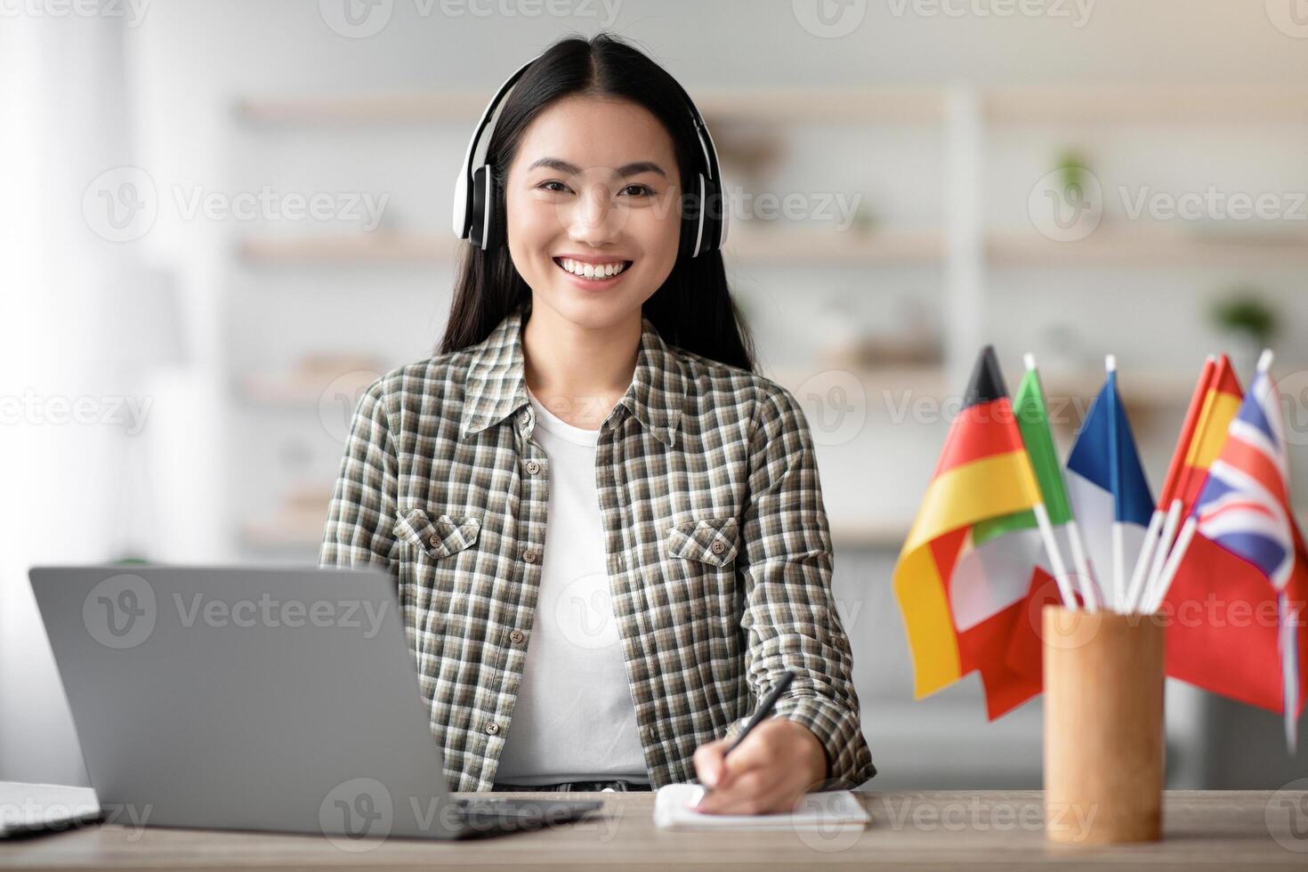 Smiling asian lady student using laptop and headset, learning foreign language online, looking at camera, taking notes while having chat with tutor, living room interior, copy space photo